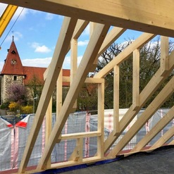 Bau eines Holzgerüsts bei Zimmereiarbeiten vor dem Hintergrund einer mittelalterlichen Kirche mit Ziegeldach und Turmuhr.