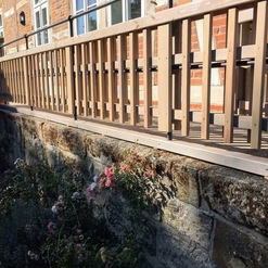 Neu installiertes Holzgeländer entlang einer verwitterten Steinmauer vor einem historischen Backsteinhaus.