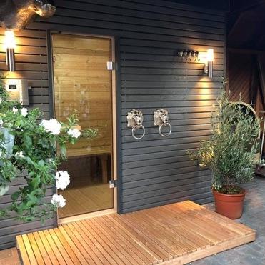 Moderner Saunabau mit dunklen Holzwänden, beleuchtet von Wandlampen. Ein Holzdeck führt zur Glastür. Dekoriert mit blühenden Pflanzen und zwei metallischen Türklopfern in Elefantenform.