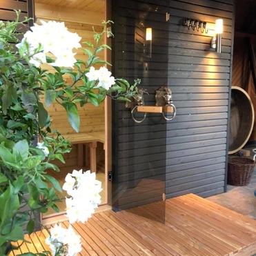 Stilvolle Außensauna mit dunkler Holzfassade und gläserner Tür, umgeben von blühenden Pflanzen, zeigt Saunabau.