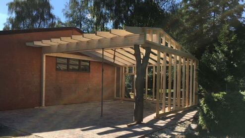 Konstruktion eines Carports aus Holz mit einem teilweise fertiggestellten Dach, nahe an einem gemauerten Gebäude.