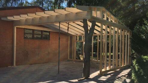 Konstruktion eines Carports aus Holz mit einem teilweise fertiggestellten Dach, nahe an einem gemauerten Gebäude.
