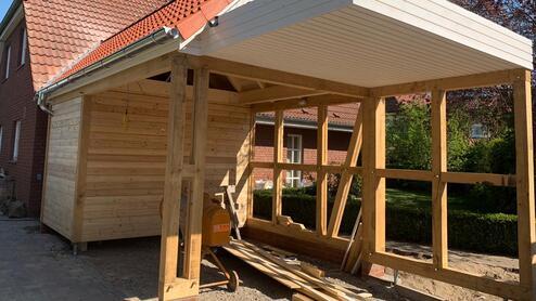 Bau eines neuen Carports neben einem Wohnhaus Konstruktion eines Carports aus Holz, halbfertig, mit sichtbarer Dachkonstruktion und roten Dachziegeln, im Hintergrund ein Wohnhaus.