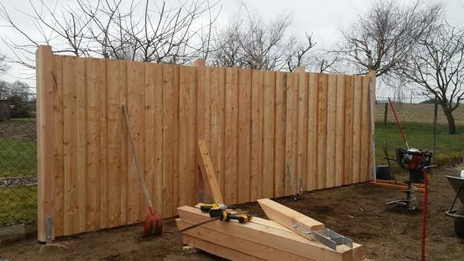 Aufbau eines hohen Holzzauns im Garten, zeigt Werkzeuge und Materialien für den Zaunbau auf einem frisch bearbeiteten Boden.